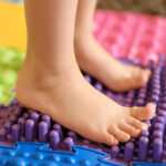 Baby foot standing on an orthopedic massage mat made of puzzles with surfaces of various shapes and tactility.  grass, cones, acorns - the texture of details. Prevention and treatment of flat feet