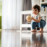 Cute little playful girl child learning to walk and stand by taking support of couch at home while looking away mischievously