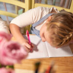 From above of blond girl sitting at table and writing in copybook while doing exercise in classroom