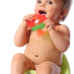 Pretty happy baby child sits on green potty and holds teether in hands. Baby loved toy. Potty training. Portrait on isolated white background